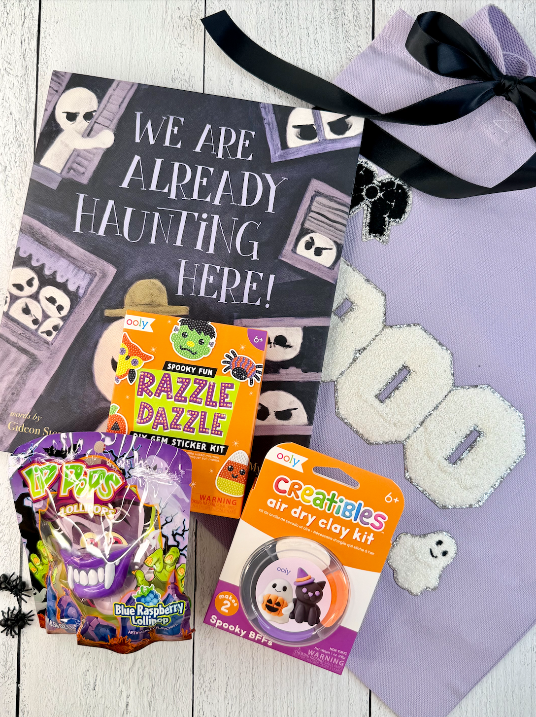 A Halloween-themed gift basket or bundle laid out on a white wood surface, including a large black sign that reads 'WE ARE ALREADY HAUNTING HERE!', a purple bag of Bling Bling Raspberry Ice pops, a Funko Razzle Dazzle sticker kit, a Creatives air dry clay kit, and a lavender ghost-themed bag with a black ribbon.
