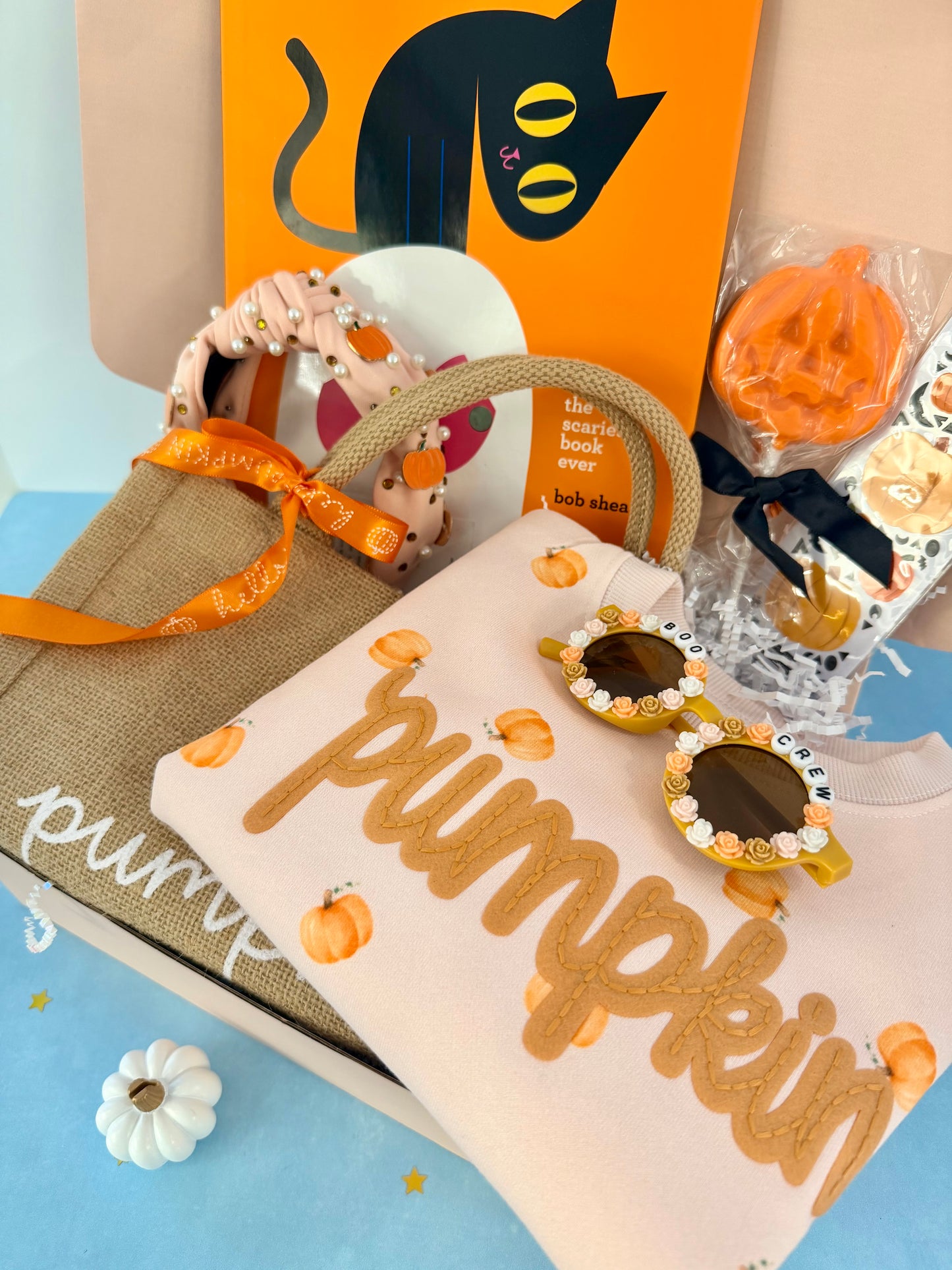 A styled gift box arrangement featuring a beige tote bag with an orange ribbon, a cream sweatshirt printed with 'pumpkin' in tan letters and small pumpkin motifs, novelty sunglasses with jeweled frames, a small pumpkin ornament, and an orange book or card with a black cat illustration.