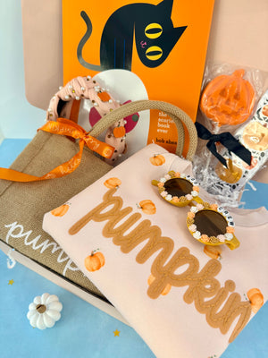 A styled gift box arrangement featuring a beige tote bag with an orange ribbon, a cream sweatshirt printed with 'pumpkin' in tan letters and small pumpkin motifs, novelty sunglasses with jeweled frames, a small pumpkin ornament, and an orange book or card with a black cat illustration.