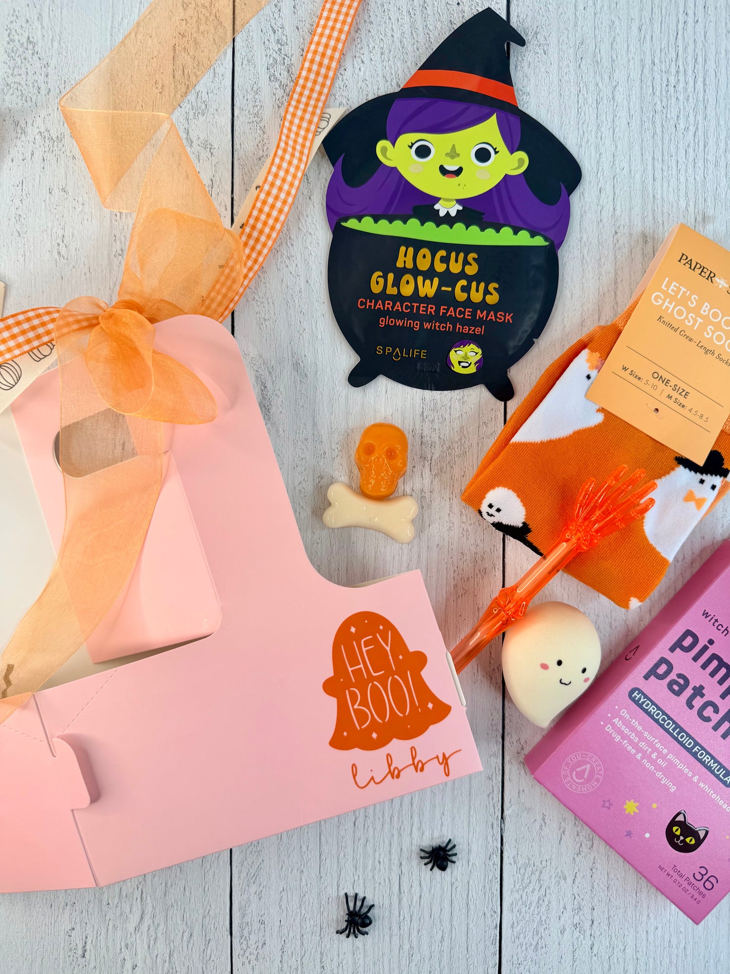 A styled flat-lay of Halloween-themed beauty products on a whitewashed wood background, including a pink 'Hey Boo!' box, a witch face mask packet, orange gummy candy, a ghost sock, and a pink patch box.