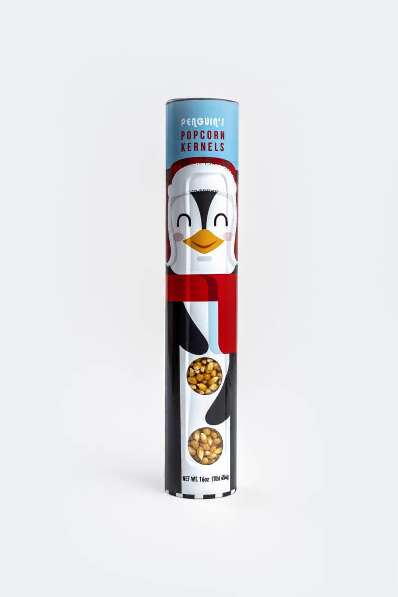 A tall cylindrical package of popcorn kernels featuring a cartoon penguin design with a light blue top section, red scarf, black-and-white body, and two clear windows showing the yellow popcorn kernels inside, photographed on a light gray background.