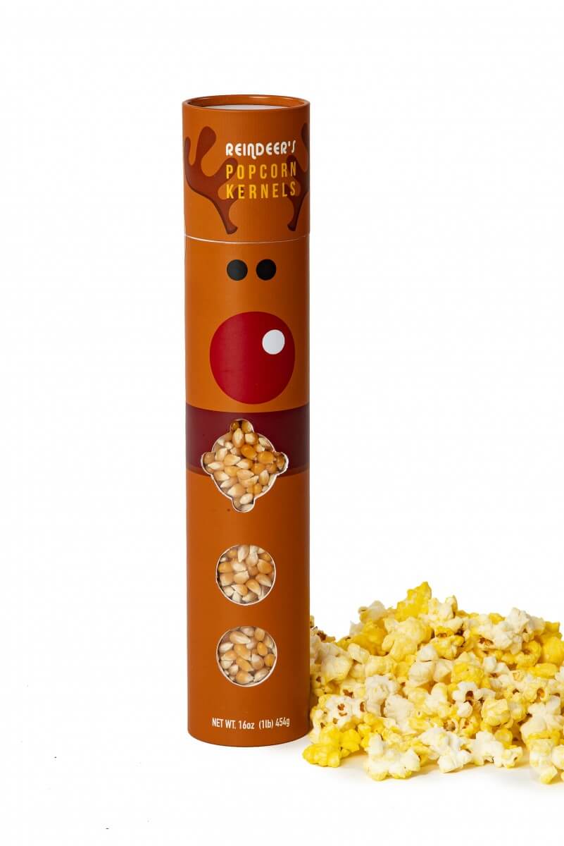 A cylindrical brown package labeled 'Reindeer Popcorn Kernels' stands on a white background with a pile of yellow and white popcorn kernels beside it. The package has a reindeer face graphic and three oval windows showing the popcorn kernels inside.