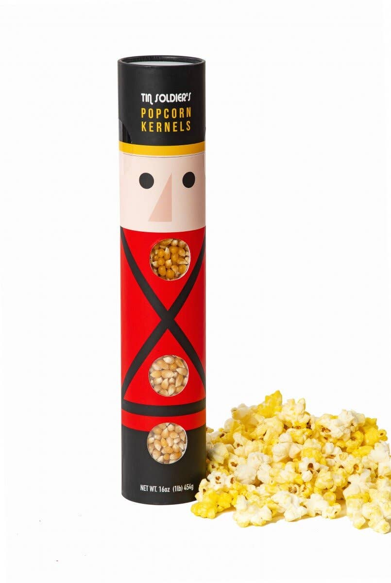 A cylindrical container of Tr. Folchey's Popcorn Kernels featuring a red and black soldier design, shown next to a pile of yellow and white popcorn kernels on a white background.