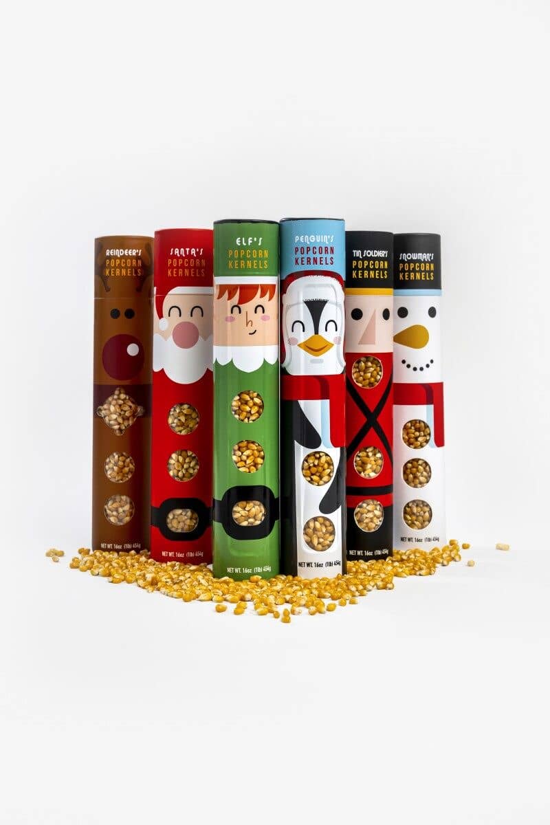 Six colorful cylindrical snack tubes standing upright on a white background with scattered golden popcorn kernels at the base. The tubes feature holiday character designs: a bear, Santa, Elf, penguin, and snowman, each with a circular window showing the popcorn inside.