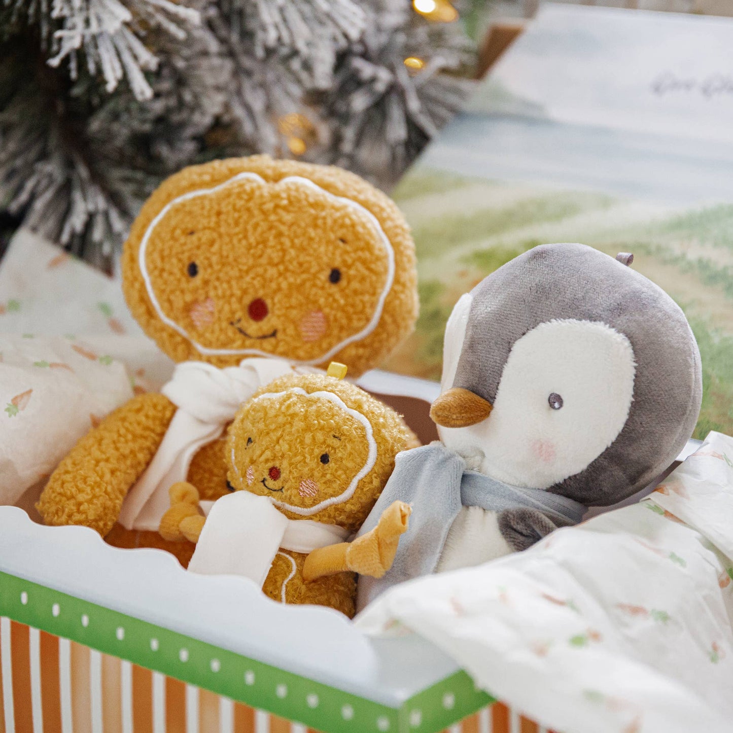 Two plush toys, a gingerbread man and a penguin, in a decorative box with a Christmas tree in the background.