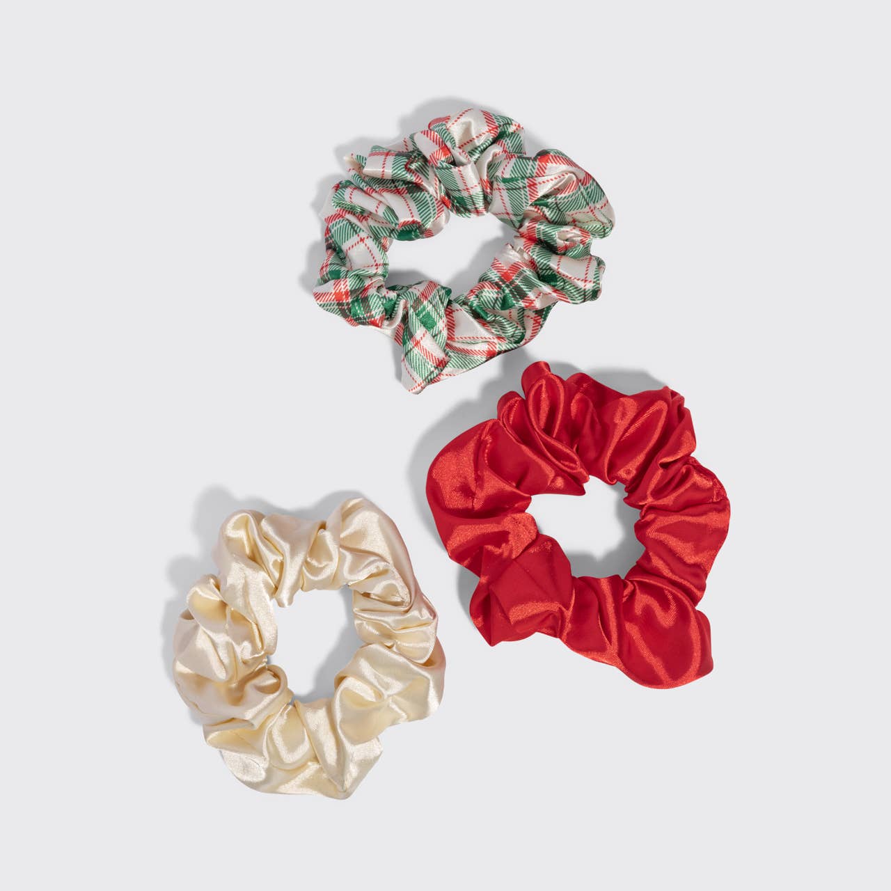 Three fabric hair scrunchies on a light gray background: one cream satin scrunchie, one red satin scrunchie, and one green and red plaid scrunchie.