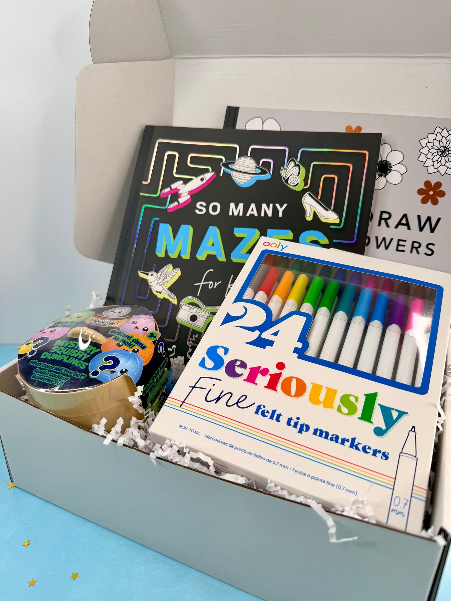 An open gift box filled with stationery and book-like items, including a box of 24 Seriously Fine felt tip markers in the foreground, a black book titled 'So Many Mazes for Her,' and a floral drawing book in the background.