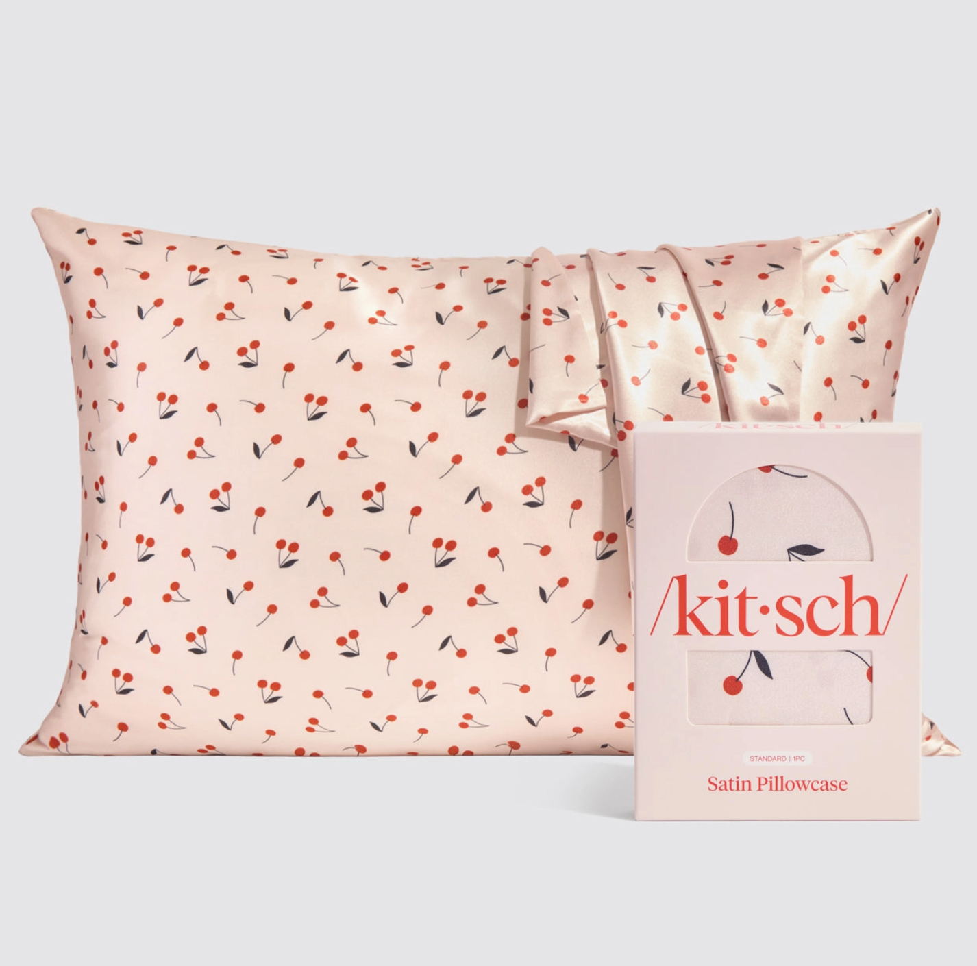 A satin pillowcase with a cherry print pattern, displayed alongside its packaging box with the brand name 'kitsch' on it.