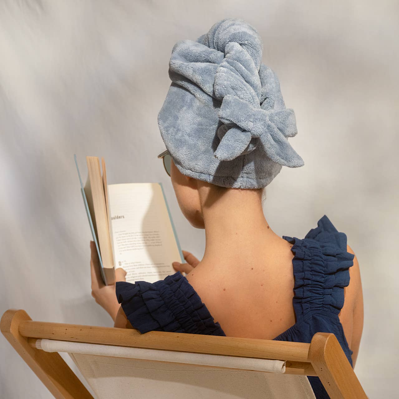 Person sitting in a chair with a blue towel wrapped around their head, reading a book.