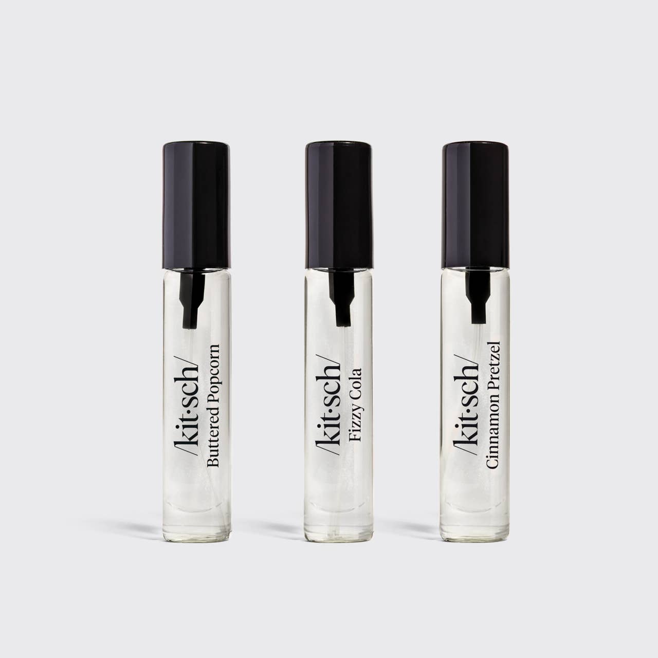 Three clear glass roll-on perfume bottles with black caps, labeled Kit's Cheddar Buttered Popcorn, Kit's Cheddar Fizzy Cola, and Kit's Cheddar Cinnamon Pretzel, arranged on a light gray background.