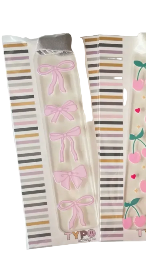 Three decorative acrylic nail strips in their original packaging, featuring pink bows, pink cherries with green stems, and red cherries with pink bows, all on striped backing cards.