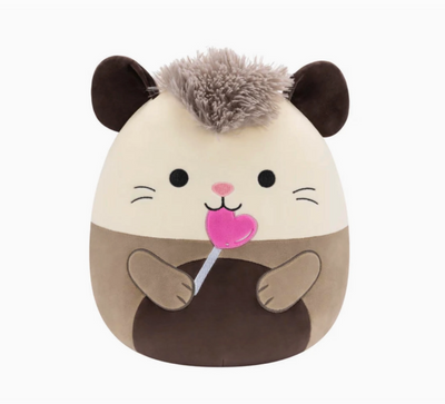 A beige and brown Squishmallow plush toy in the shape of an opossum, holding a lollipop, with a soft, marshmallow-like texture.