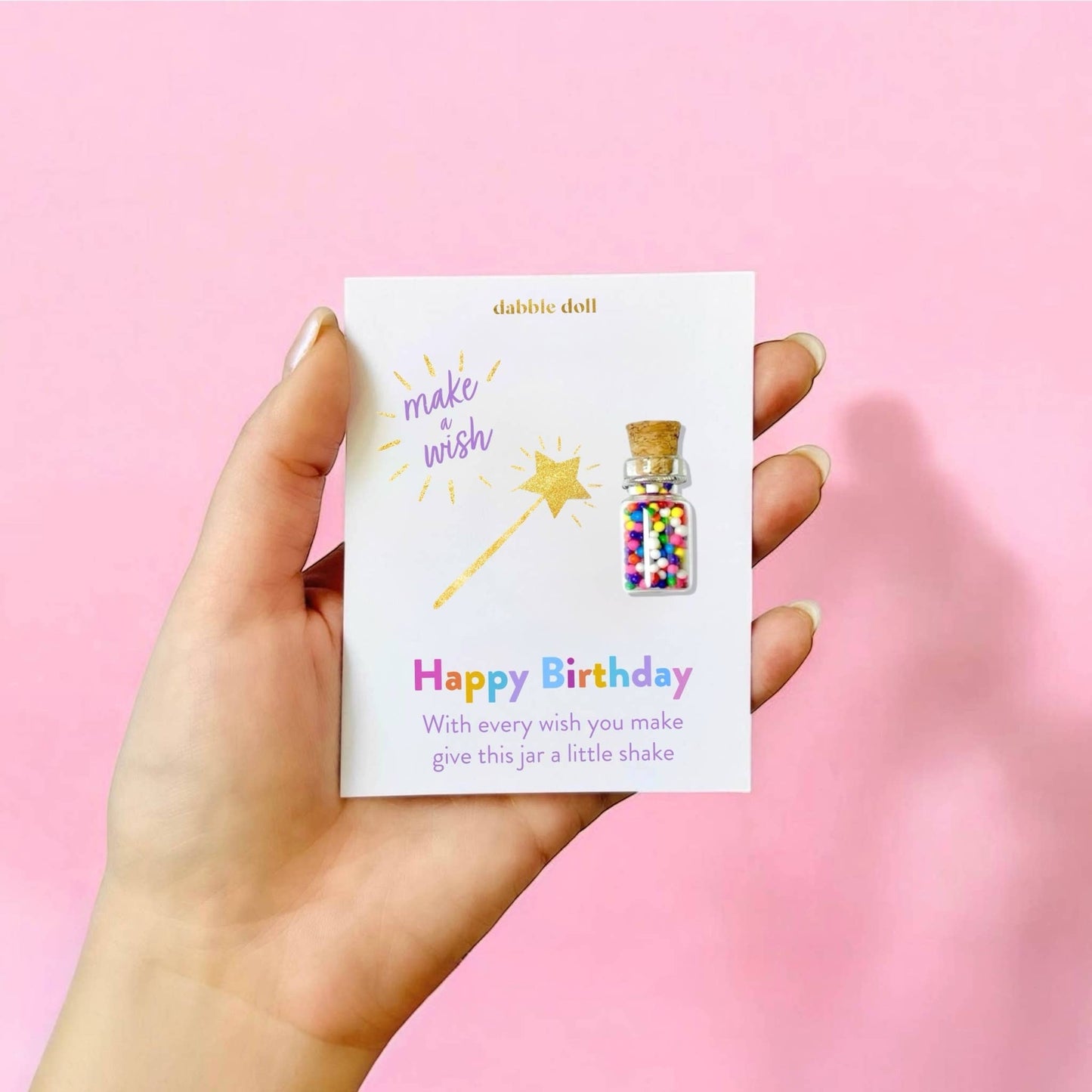 A hand holds a white card featuring a small birthday gift: a glittery bottle pin with colorful confetti and a gold star wand illustration. The card reads 'dabble doll' and 'Happy Birthday'.