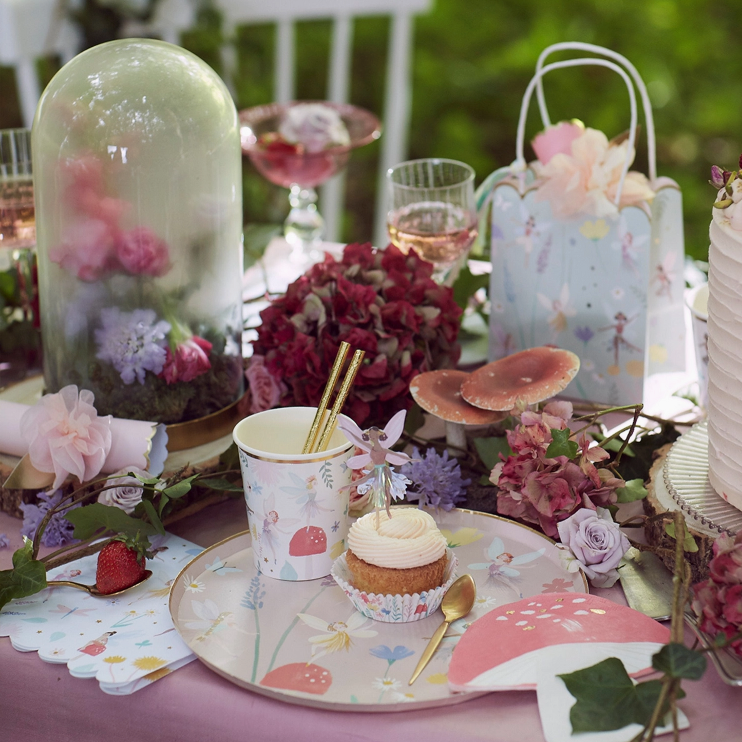 Table setting with fairy-themed decor, including a cupcake, flowers, and decorative items on a pink tablecloth.