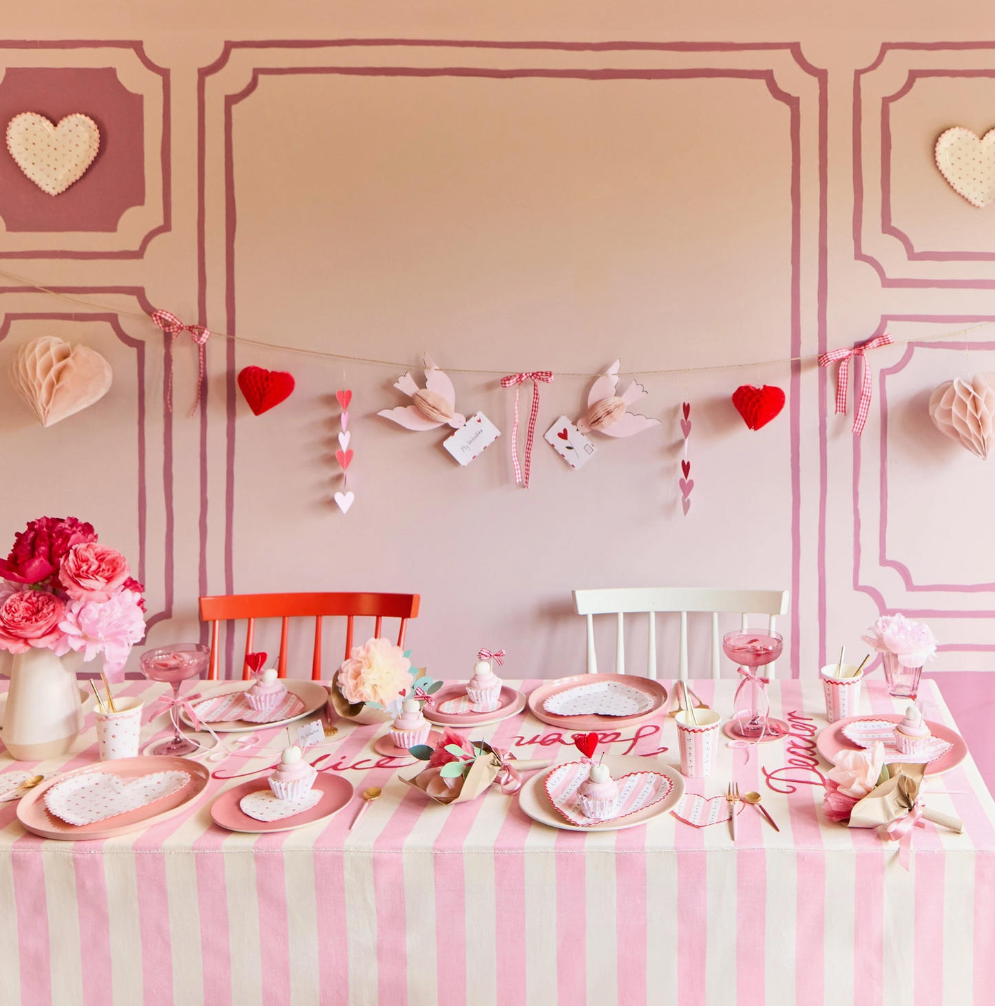 Children's Valentine's Day table setting with pink and white striped tablecloth, heart decorations, and floral arrangements.