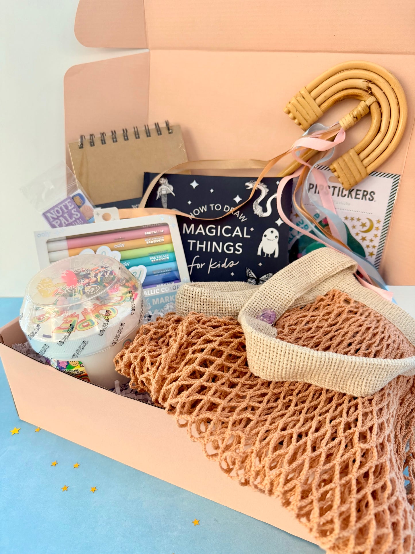 A pastel pink gift box filled with handmade and school-supplies items, including a woven tan mesh bag, a wooden hula hoop, a notebook, stickers, and a book titled 'How to Do Magical Things for Kids'.