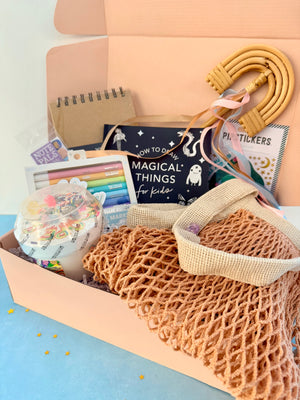 A pastel pink gift box filled with handmade and school-supplies items, including a woven tan mesh bag, a wooden hula hoop, a notebook, stickers, and a book titled 'How to Do Magical Things for Kids'.