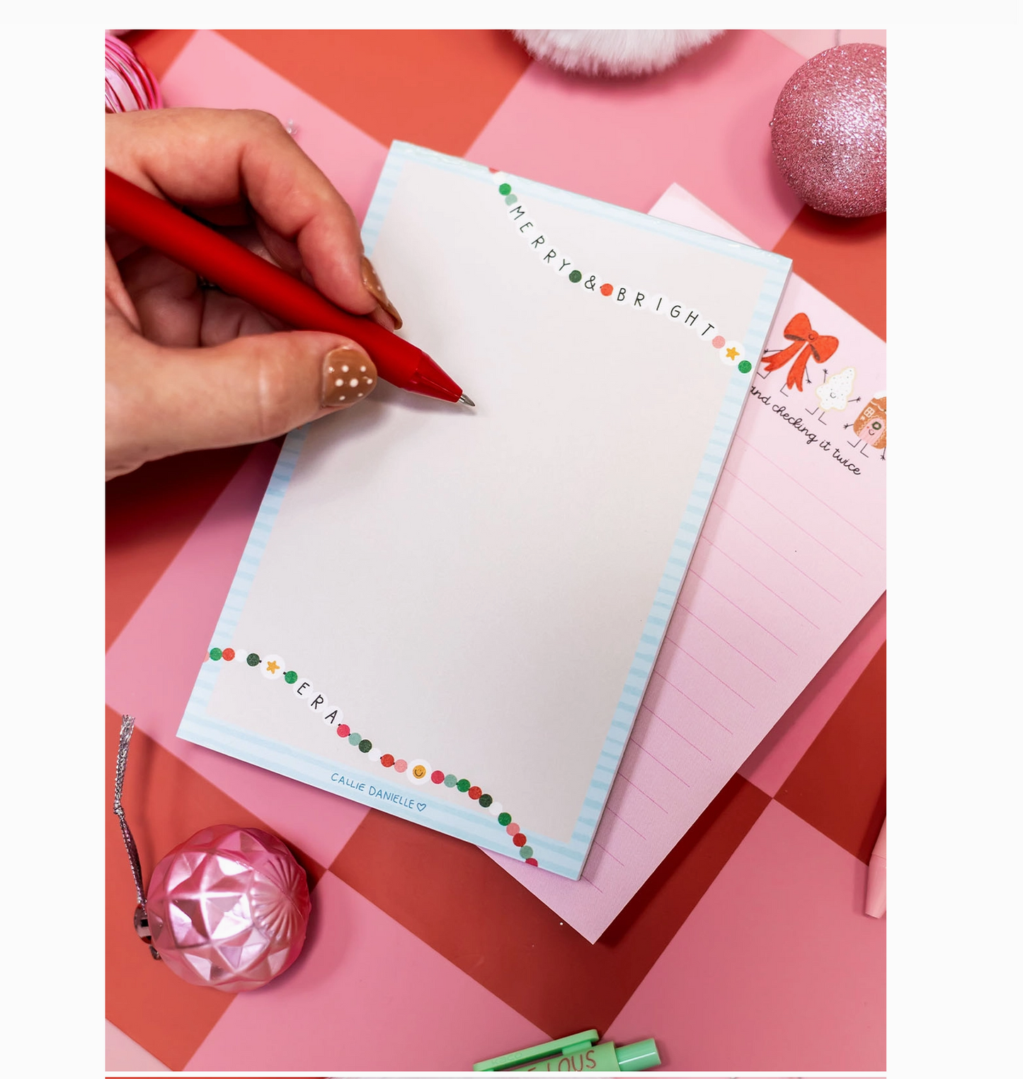 Hand writing on a Christmas-themed notepad with decorative items on a pink and red checkered background.