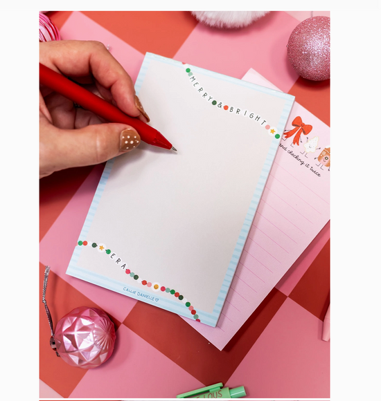 Hand writing on a Christmas-themed notepad with decorative items on a pink and red checkered background.