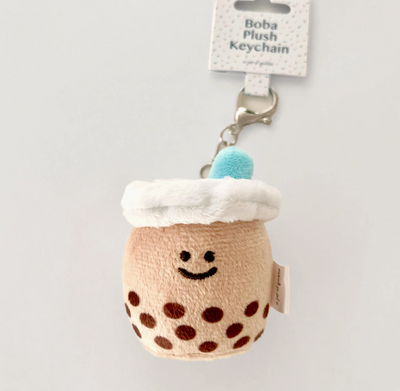 A small plush keychain in the shape of a boba milk tea cup with a white and brown color scheme, featuring a smiling face, a white pom-pom on top, and a blue-patterned surface.