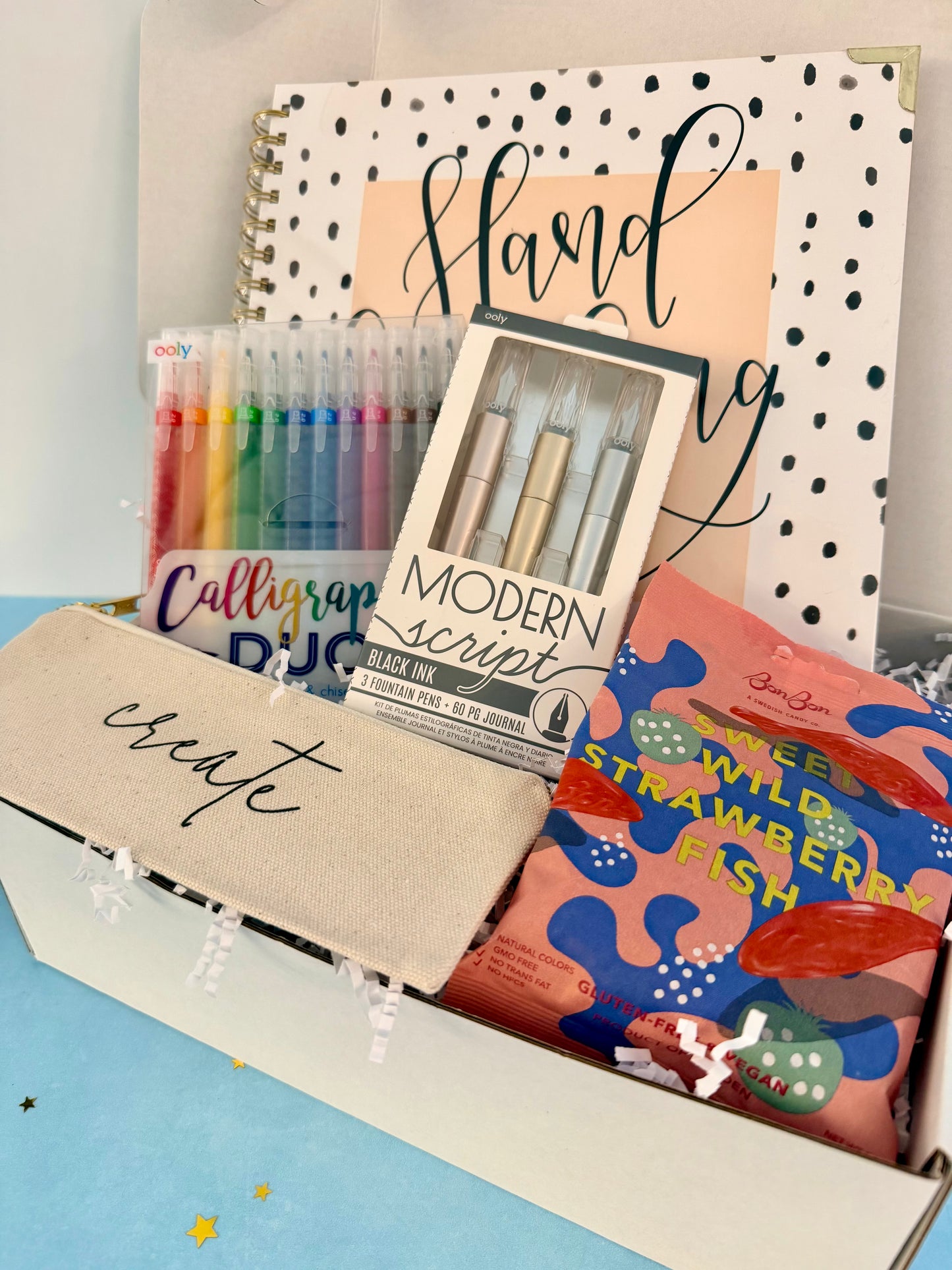 A curated stationery gift box filled with calligraphy supplies and snacks. Visible items include a beige fabric pouch with 'create' in script, a box of Modern Script pens, a set of colorful calligraphy pencils, a notebook, and a bag of Sweet Wild Strawberry Fish.