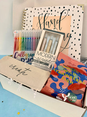 A curated stationery gift box filled with calligraphy supplies and snacks. Visible items include a beige fabric pouch with 'create' in script, a box of Modern Script pens, a set of colorful calligraphy pencils, a notebook, and a bag of Sweet Wild Strawberry Fish.