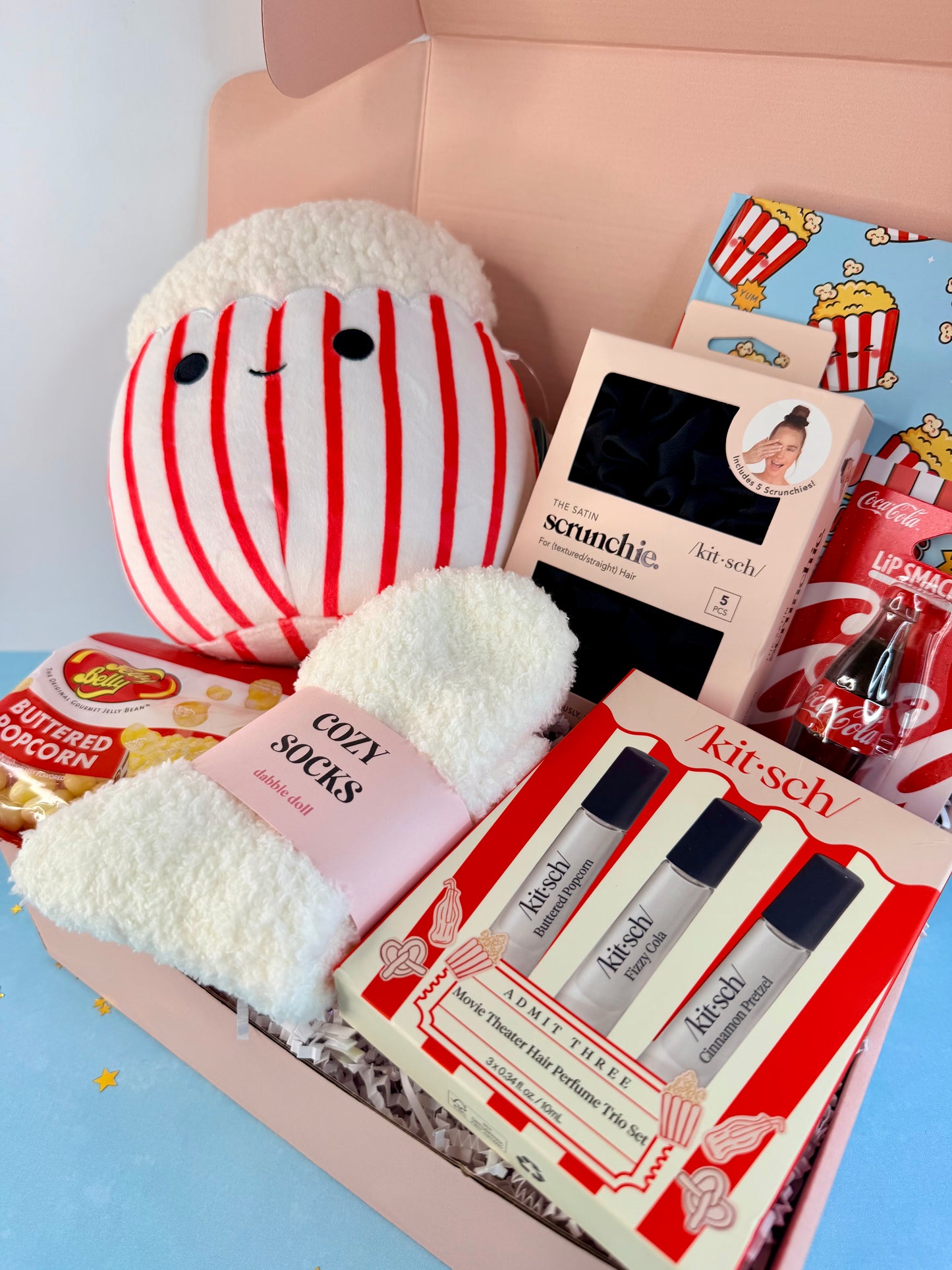 A curated gift box filled with assorted beauty and snack items, including a red-and-white striped plush toy, fluffy white socks, a boxed scent kit labeled 'kitsch', two small perfume-style bottles, candy bars, and popcorn-themed packaging, all arranged in a pale pink box.