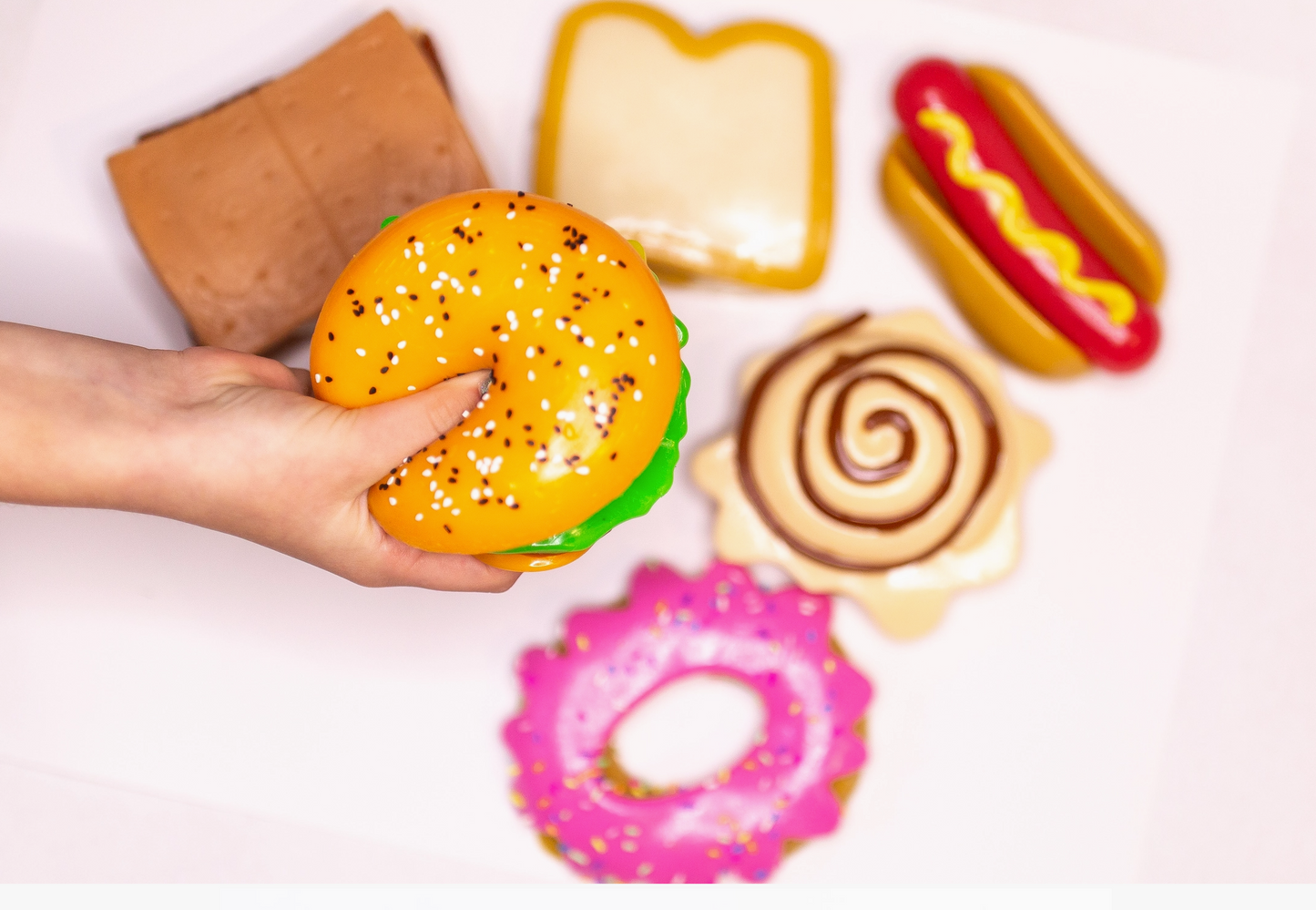 Hand holding a toy hamburger with other food-themed toys on a light background