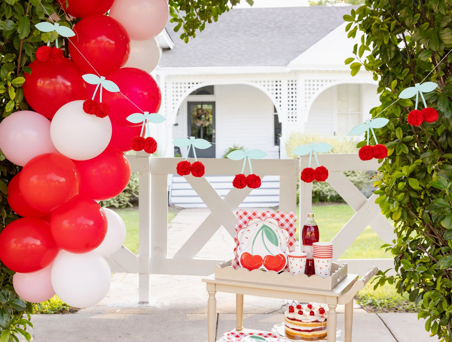 Outdoor setting with red, white, and pink balloons, cherry decorations, and a small table with a cake.