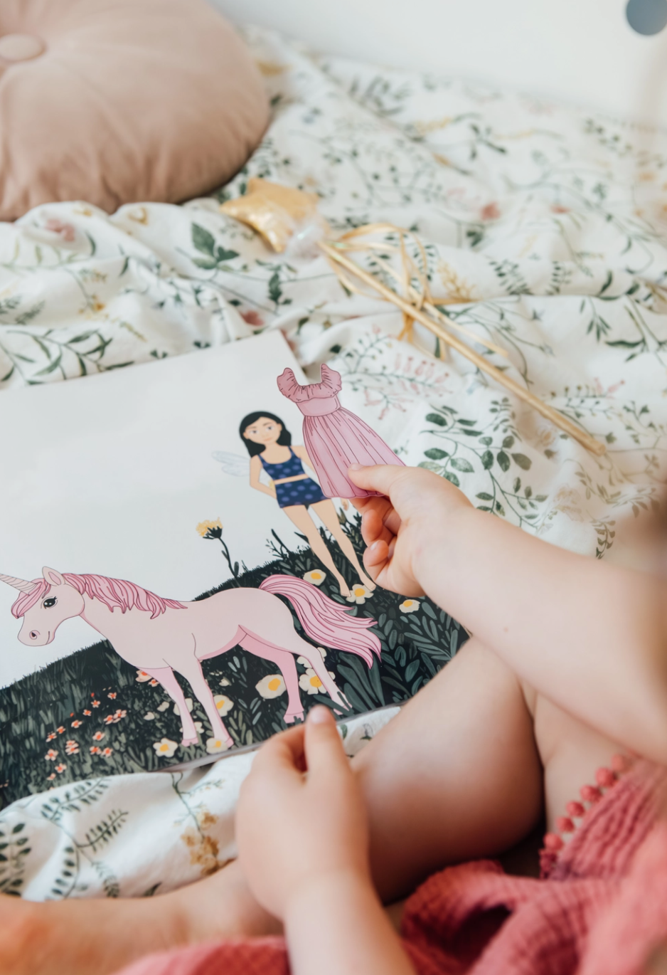 Child's hand holding a book with a unicorn illustration on a bed with floral bedding.