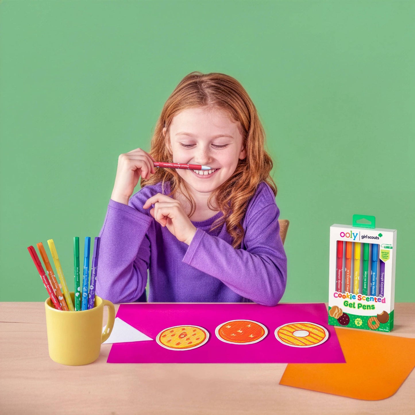 Girl playing with colorful cookie-shaped gel pens on a table against a green background