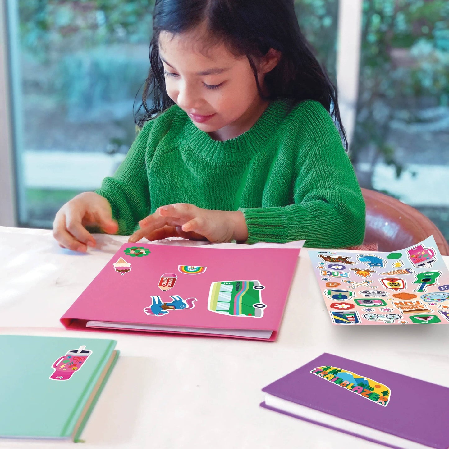 Child in a green sweater reading a pink book with colorful illustrations on a table.