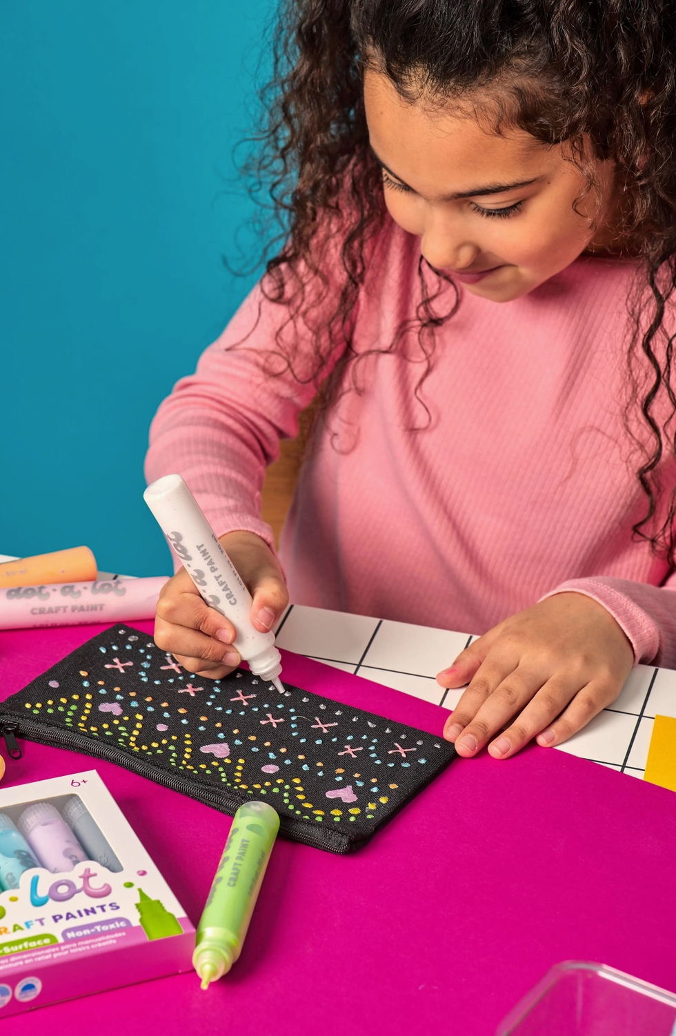 Child painting on a black canvas with markers, sitting at a table against a colorful background.