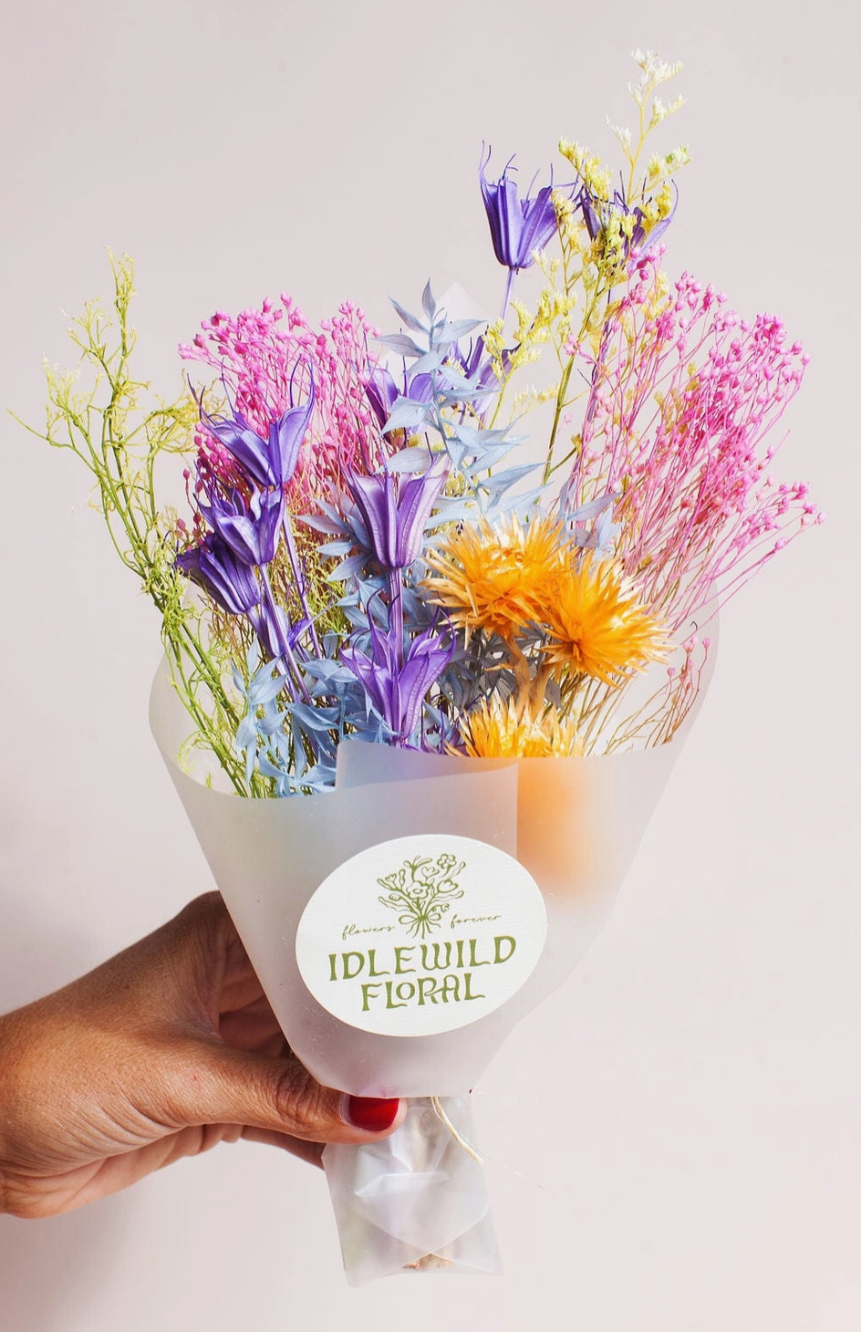 Bouquet of colorful flowers held by a hand with 'Idel Wild Floral' label on a light background