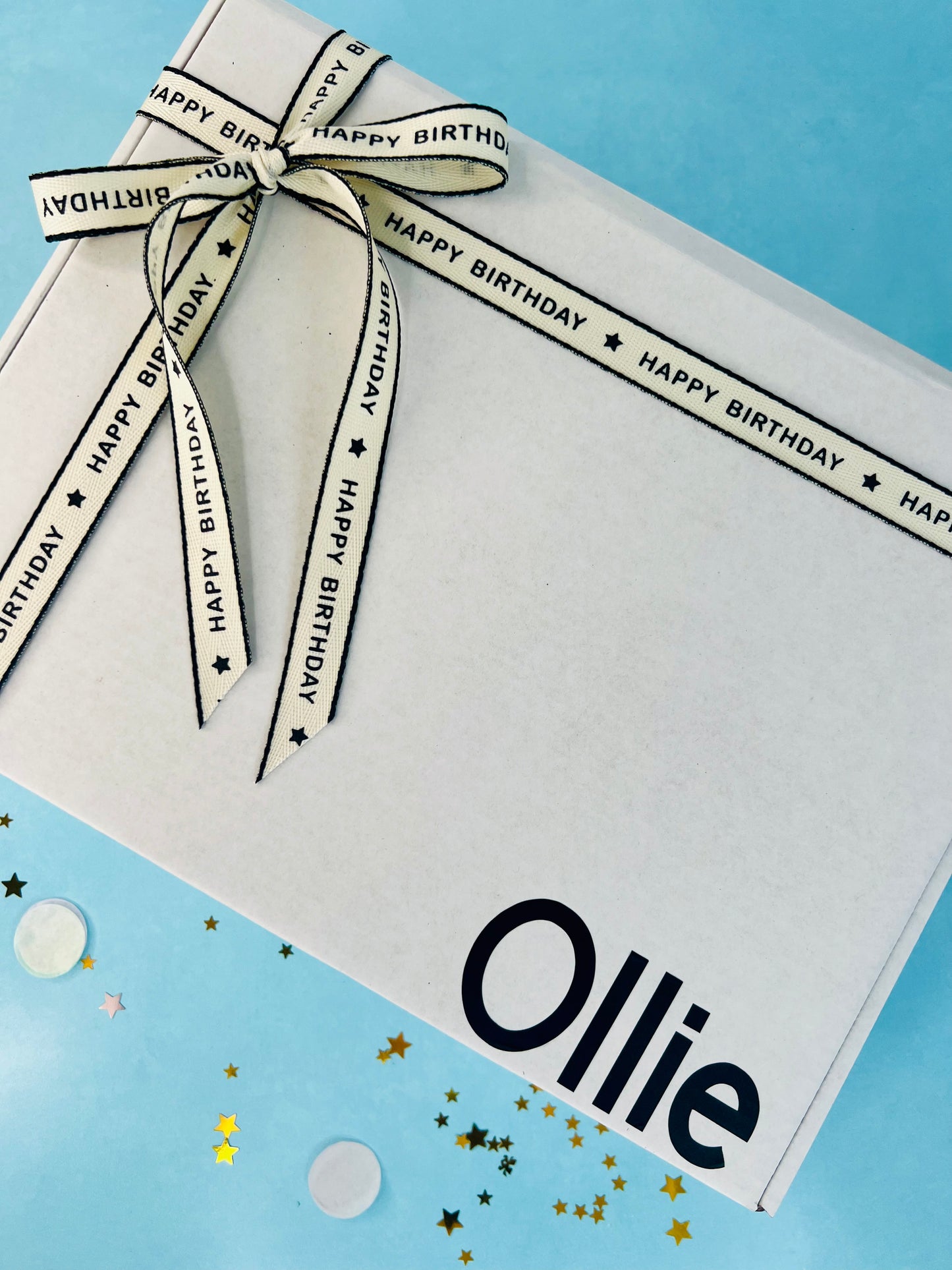A white and black gift box with 'Ollie' written on it, adorned with a decorative ribbon and tissue papers, on a blue background with confetti scattered around.