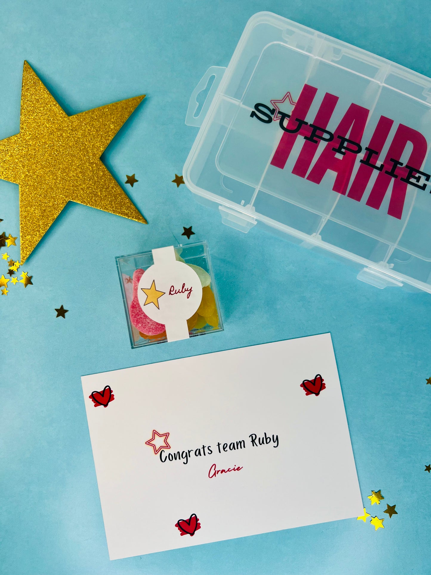 A styled flat-lay of hair supply accessories on a light blue background, including a clear plastic storage box labeled 'HAIR SUPPLIES', a small clear box with colorful hair ties, a white congratulatory card reading 'Congrats team Ruby', and a large gold glitter star.