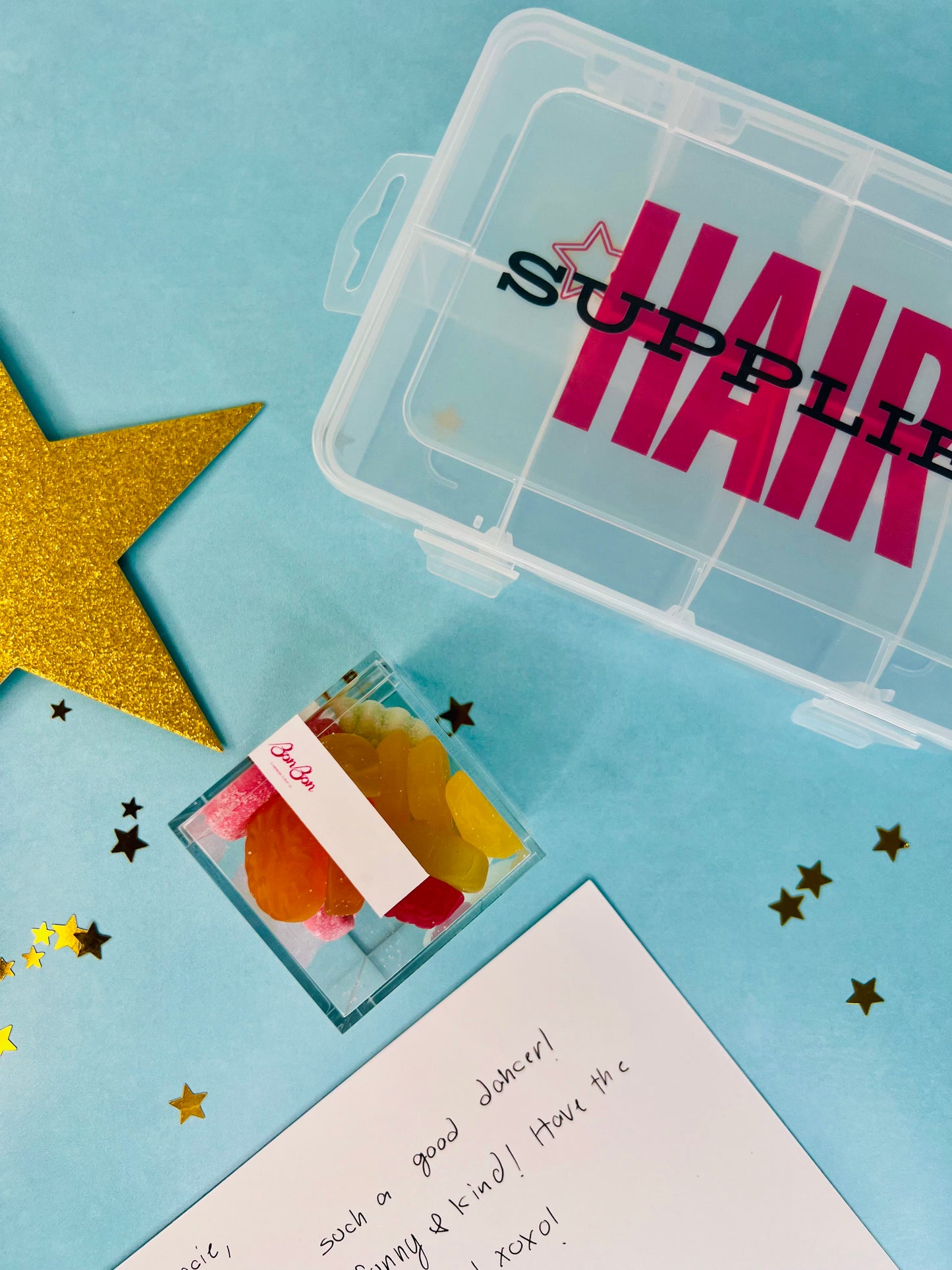 A clear plastic hair supply organizer box with pink 'HAIR SUPPLIES' text, shown alongside a small clear container of colorful hair clips or accessories on a light blue background with gold star confetti.