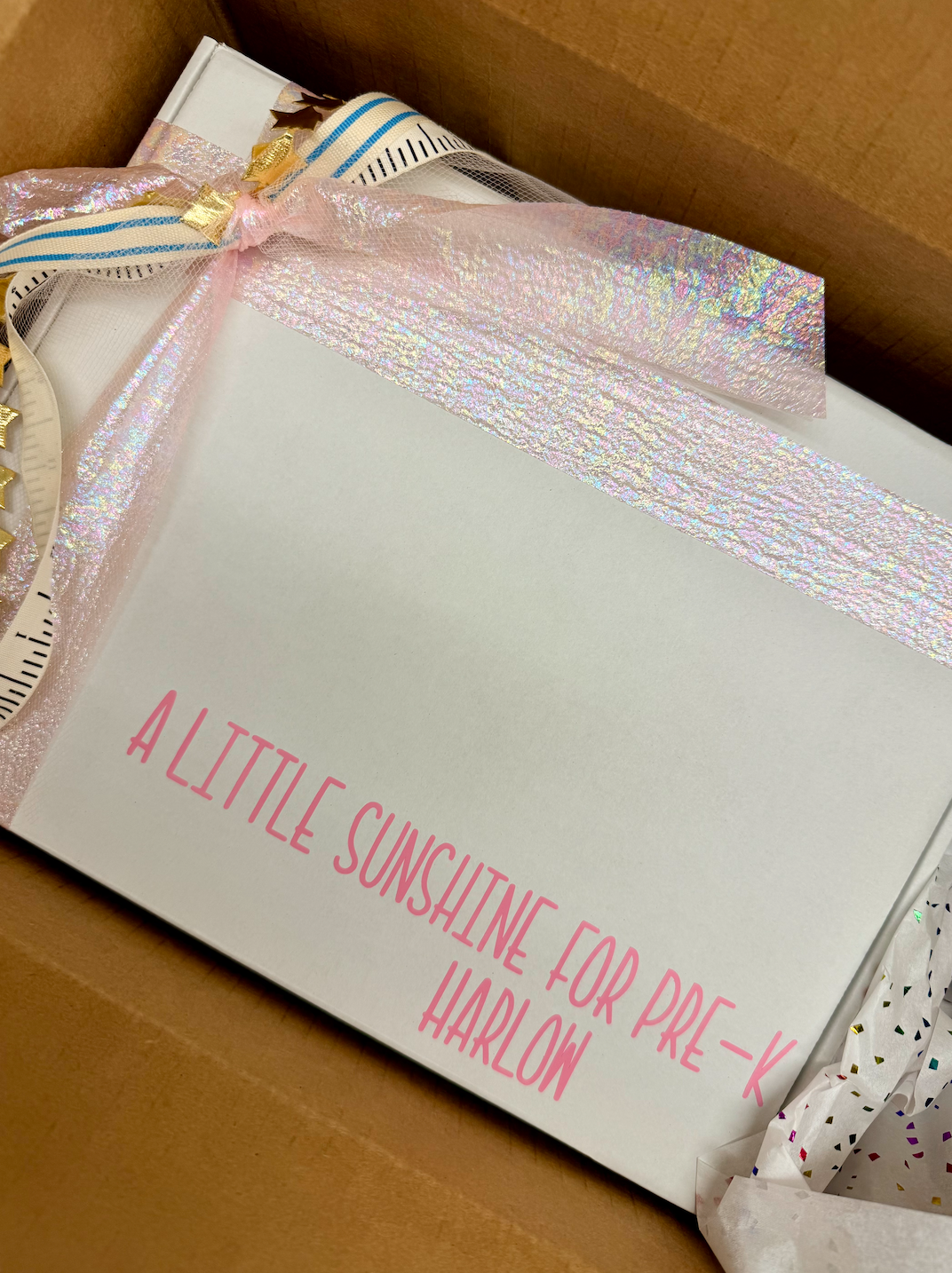 A white personalized gift box or card inside a brown cardboard box, decorated with a pink iridescent ribbon and a striped paper ribbon. Pink text on the front reads 'A LITTLE SUNSHINE FOR PRE-K HARLOW'.