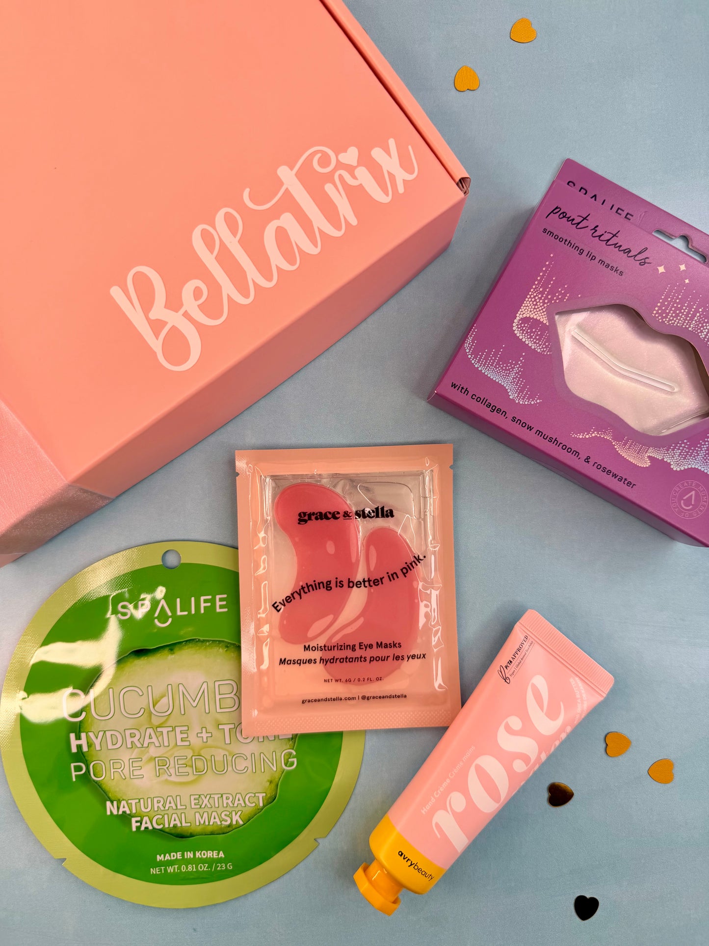 A curated beauty gift set featuring a pink Bellatrix box, a purple Naitice facial mask box, a green Tsai Life cucumber mask packet, a peach-colored Grace + Stella micellar eye mask packet, a pink tube labeled 'rose', and a peach-colored tube, all arranged on a light blue background with small heart-shaped confetti.