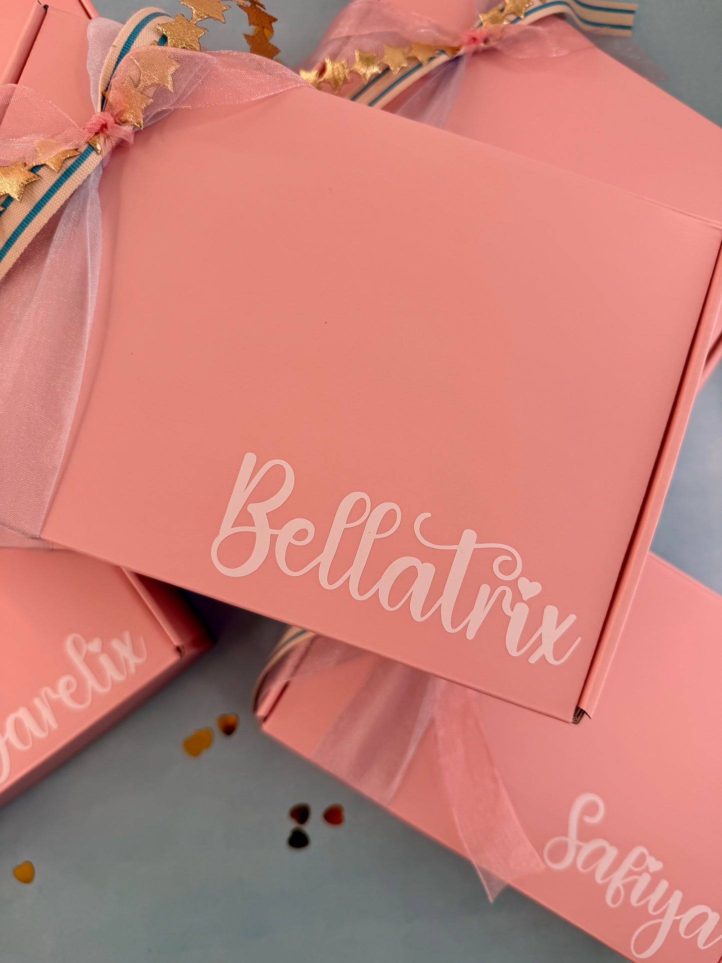 Pink gift boxes with white script branding, including Bellatrix and Safiya, tied with sheer pink ribbon and gold leaf accents on a light blue surface.