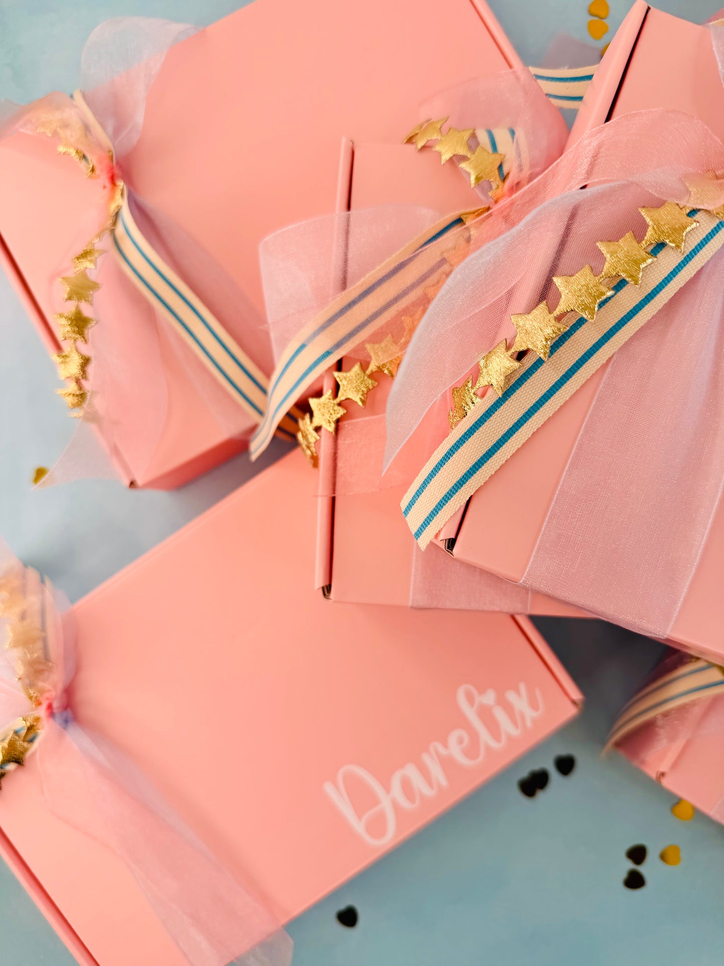 Personalized pink gift boxes with tulle ribbon, gold star garlands, and blue stripes, arranged on a light blue background. One box is printed with the name 'Darelix'.