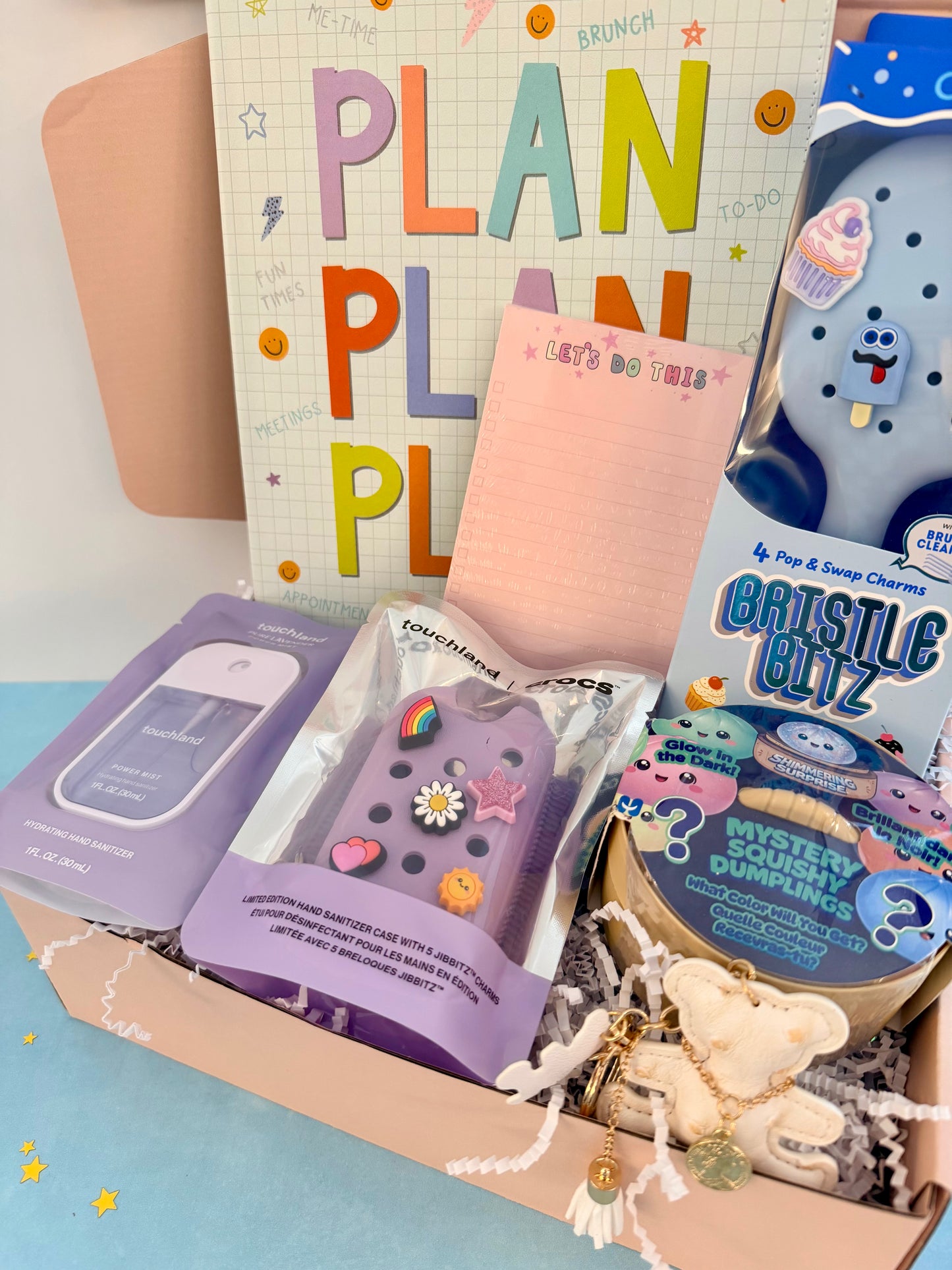 A curated gift box filled with cute stationery and small accessories, including a colorful 'PLAN PLAN' card, a pink notepad, a purple phone case, a boxed blue plush toy, and a round tin labeled 'Mystery Bubble Wrap'.