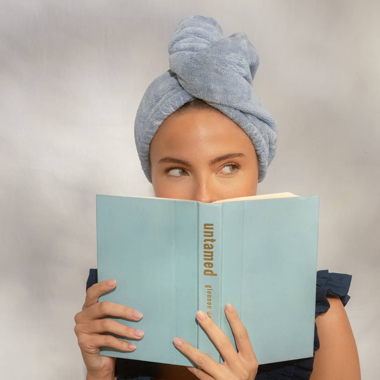 Woman with a towel wrapped around her head reading a book titled 'untamed' on a plain background