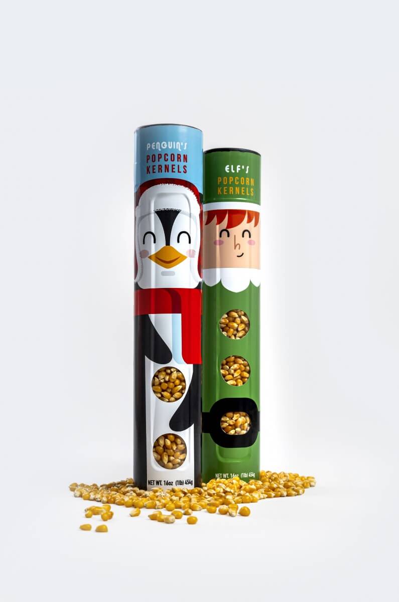 Two decorative cylindrical tubs of popcorn kernels standing upright on a white background, with loose yellow popcorn kernels scattered at their bases. The left tub is light blue with a penguin illustration and reads 'Piquadro Popcorn Kernels'; the right tub is green with an elf illustration and reads 'Elf's Popcorn Kernels'.