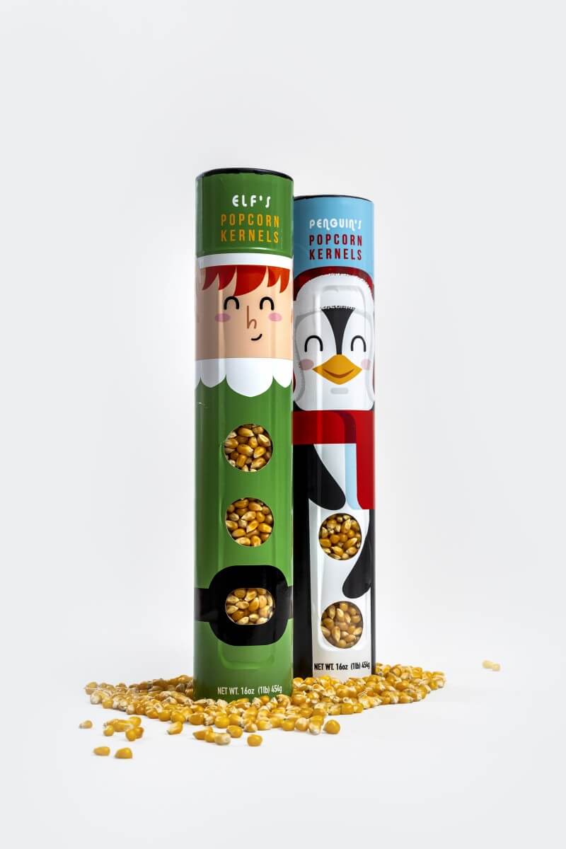 Two novelty popcorn kernels in cylindrical tubes with elf and penguin illustrations, standing upright on a white background with scattered popcorn kernels at the base.