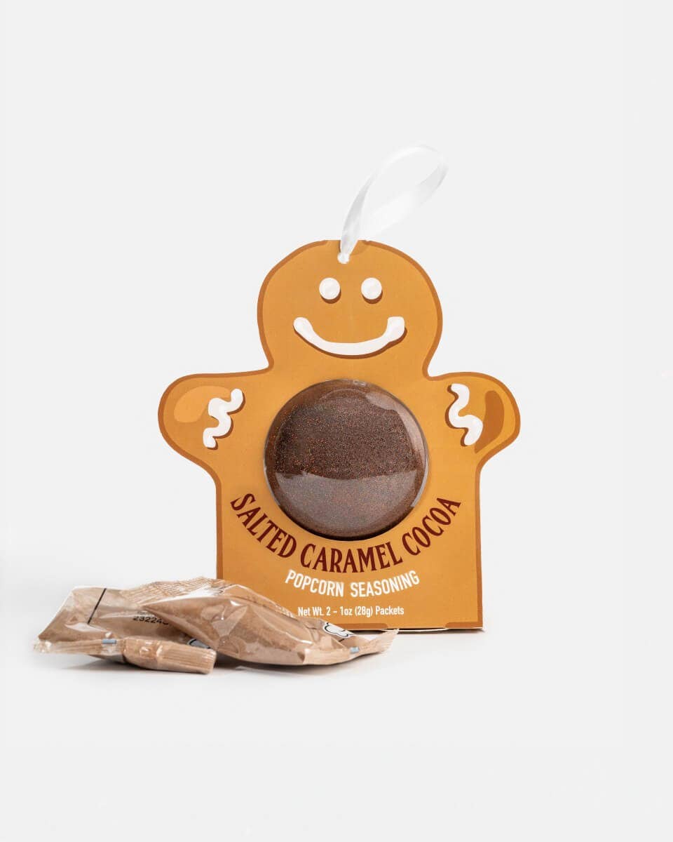 A gingerbread man-shaped container of Salted Caramel Cocoa Popcorn Seasoning with a white hanging loop, shown on a clean white background with two loose seasoning packets in front.