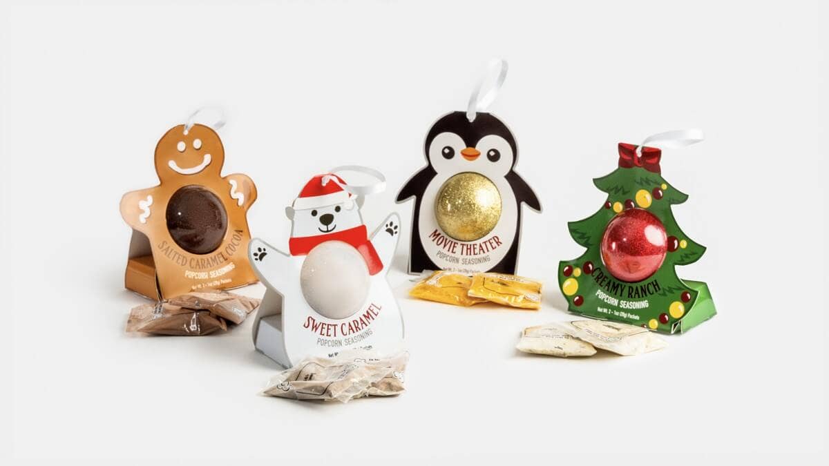 Four Christmas-themed chocolate gift boxes arranged on a white background: a gingerbread man box with a dark chocolate, a polar bear box labeled Sweet Caramel, a penguin box labeled Movie Theater with a gold foil ball, and a Christmas tree box labeled Poppy's with a red ornament.
