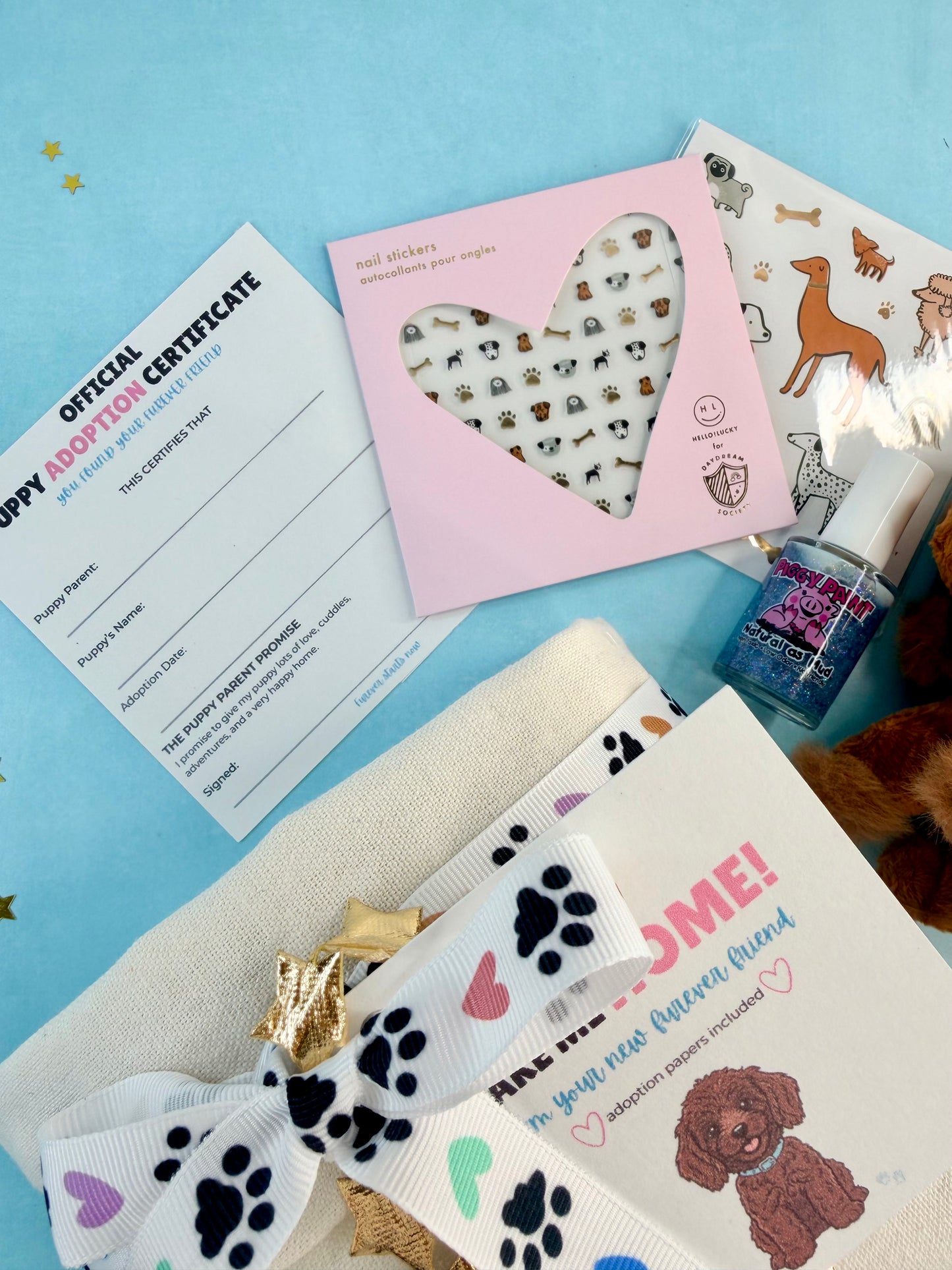 A styled flat-lay of a puppy-themed gift set on a light blue background, including a white ribbon bow with black and pastel paw prints, a pink heart-shaped nail sticker pack, a puppy adoption certificate, a small bottle of nail polish, and a printed card.