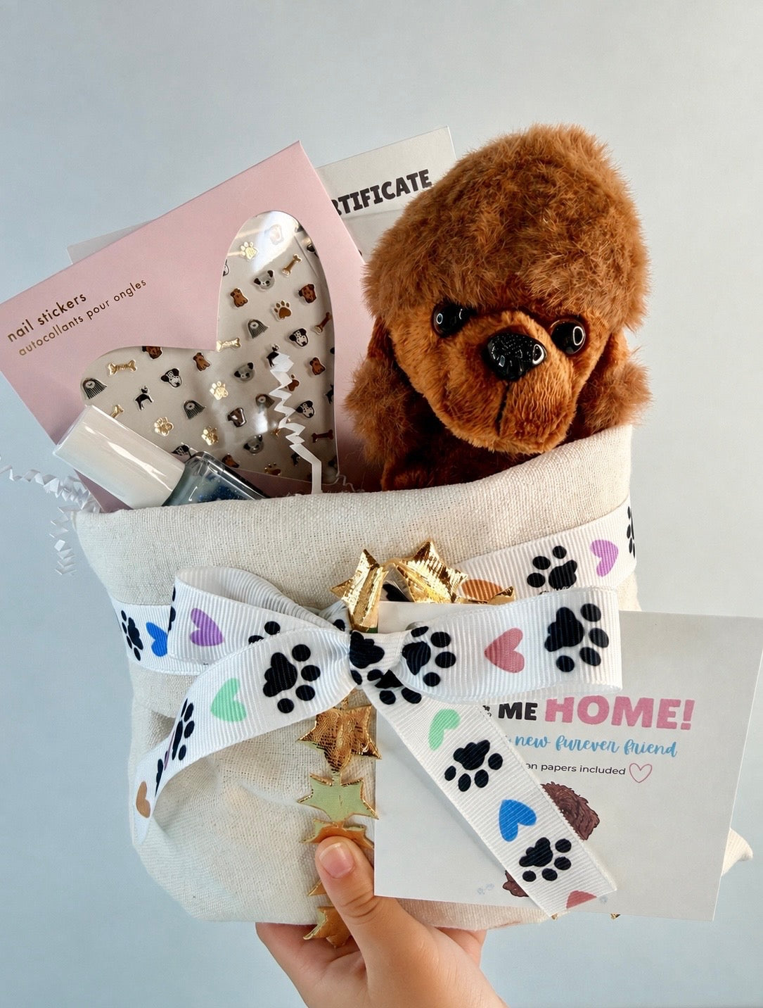 A pet memorial gift basket held by a hand, featuring a brown plush dog toy, a white fabric liner with colorful paw print and heart ribbon trim, a gold bow, nail stickers, a heart-shaped nail file, and a 'ME HOME! new forever friend' card.