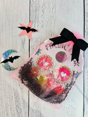 Pre-made Halloween treat bags with candy and a mask, styled in pink with black paper and ribbon, displayed on a white wooden surface with decorative star cut-outs.